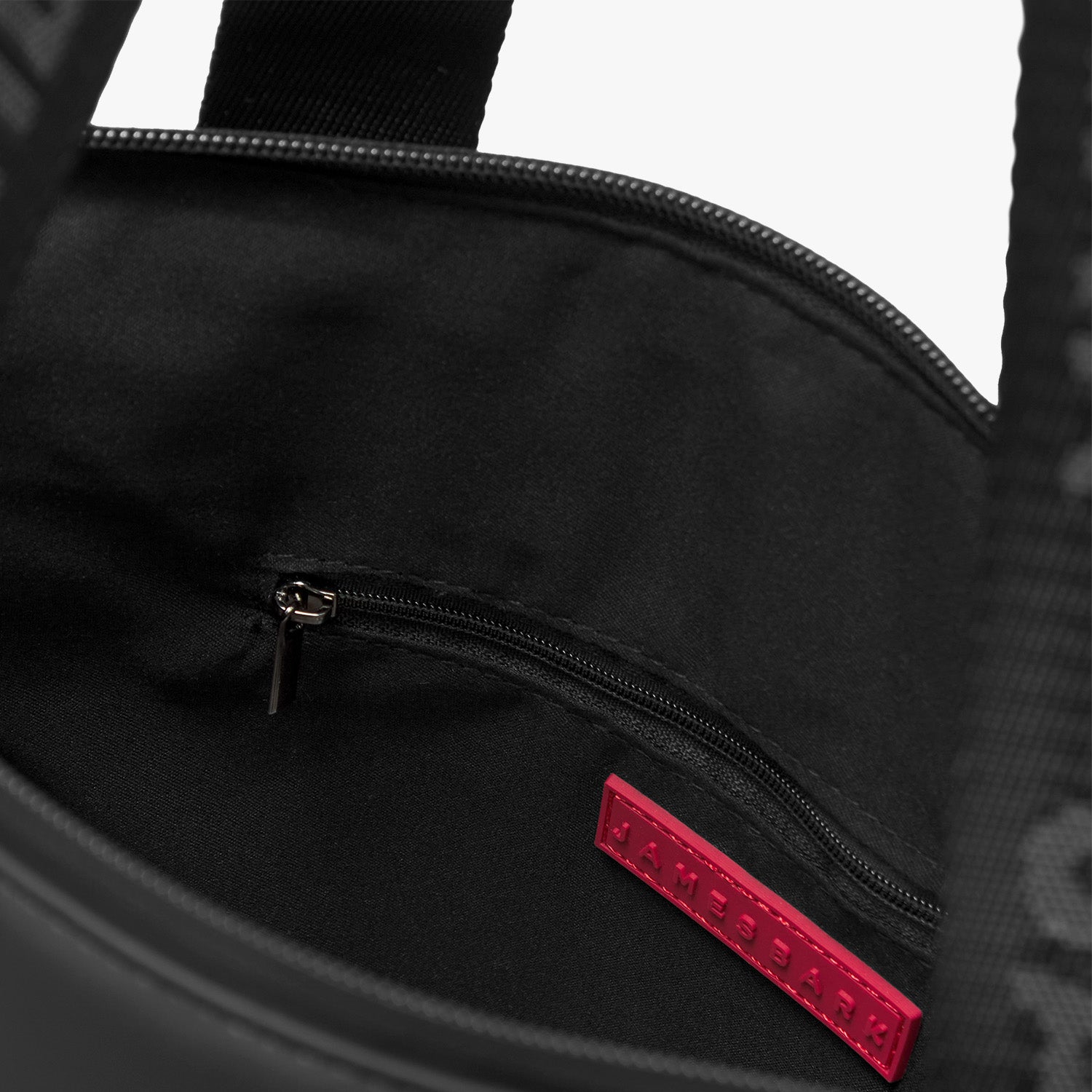 A close-up view of the interior of a black tote bag, showing its organizational features. The bag has a black fabric lining and a zippered pocket on one side for secure storage. A red rectangular brand label with the text "JAMES BARK" is stitched inside, adding a subtle contrast to the dark interior. The stitching and zipper details highlight the bag’s quality craftsmanship.