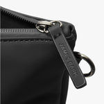 A close-up of the zipper and hardware details of a black tote bag. The zipper pull is made of black material with the brand name "JAMES BARK" embossed on it. The zipper itself is sturdy, with a smooth metal finish. A metal ring is attached near the zipper, suggesting an option for additional attachments or a shoulder strap. The stitching and matte finish of the bag highlight its sleek and durable construction.