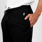 Close-up of black jogger-style sweatpants featuring an embroidered white dog logo near the pocket. The person is wearing a white T-shirt and has one hand casually placed inside the front pocket. The pants include a drawstring waist and are displayed against a plain white background." Let me know if you'd like any edits