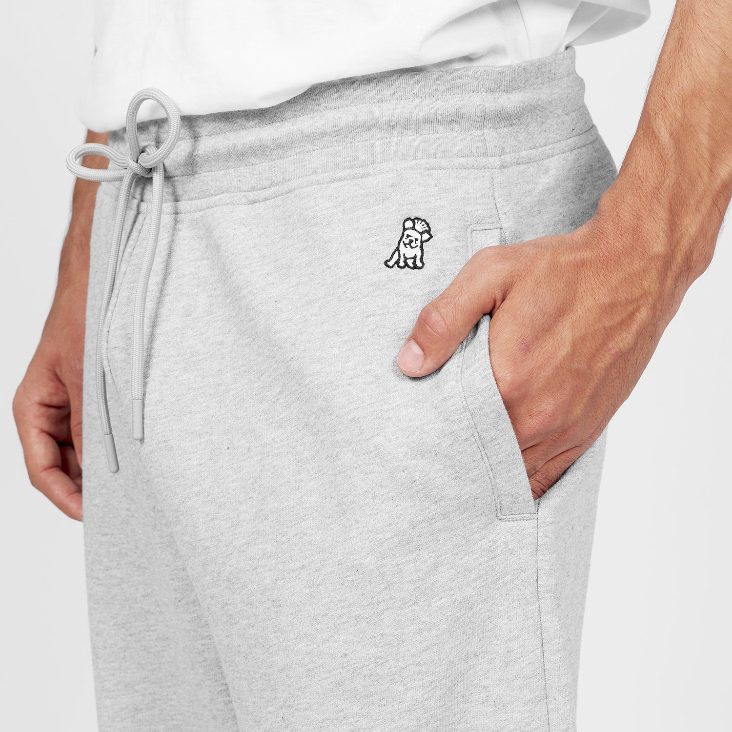 Close-up of light gray jogger-style sweatpants featuring a small embroidered dog logo near the waistband. The pants include a drawstring waist, side pockets, and a relaxed fit. A hand is placed inside one pocket, and the person is wearing a white T-shirt. The background is plain white." Let me know if you'd like any changes