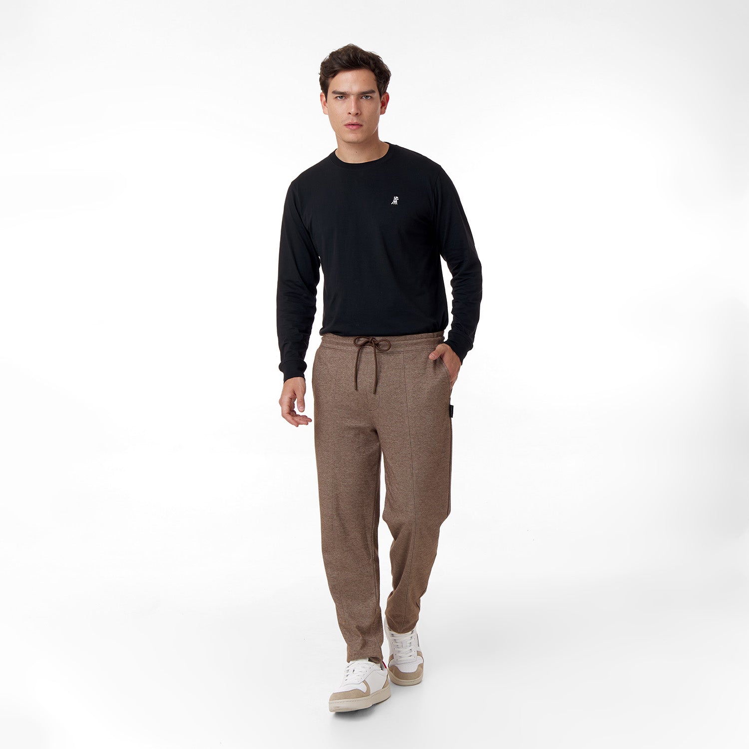 A male model wearing a black long-sleeve shirt with a small embroidered logo on the chest, paired with brown drawstring pants. He is standing in a relaxed walking pose, hands in pockets, and wearing white sneakers. The outfit is casual and stylish against a clean white background.