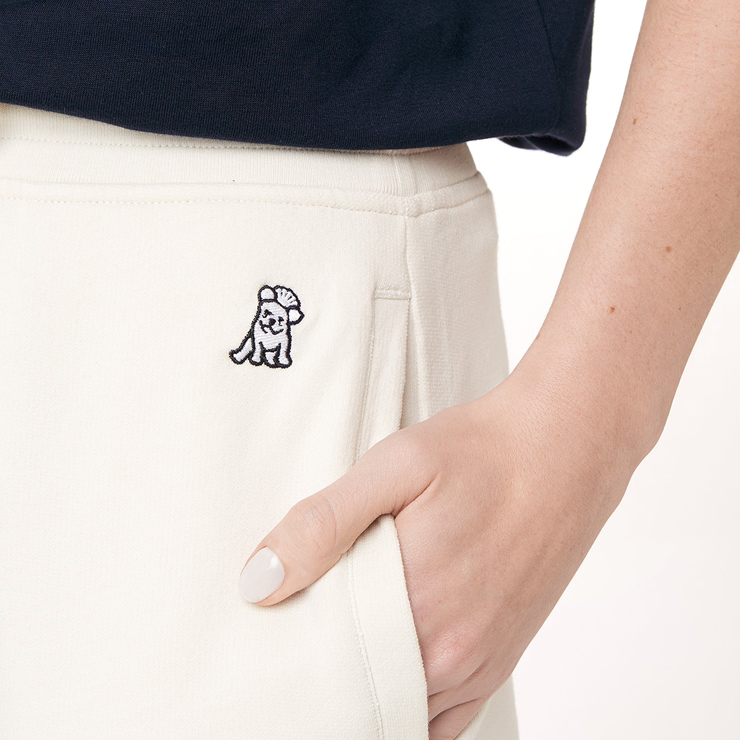 This close-up image shows a detailed view of the beige jogger pants, highlighting an embroidered logo on the left side of the waistband. The logo appears to be a small, playful design of a dog wearing a hat. The person’s hand is tucked into the pocket, and the nails are painted in a light color. The texture of the pants is also visible, emphasizing their soft, comfortable material.