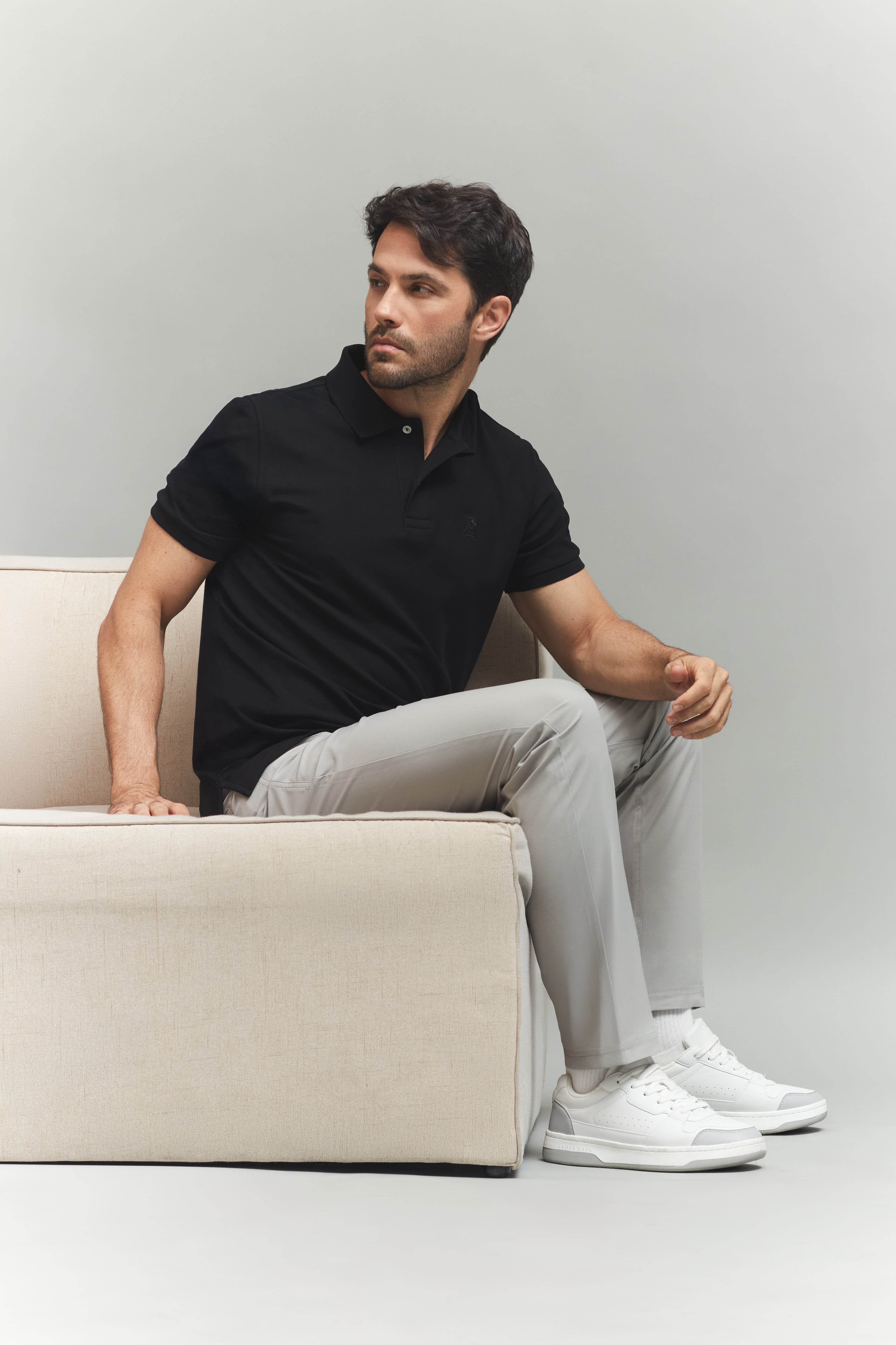 Man sitting on a beige couch wearing a black polo shirt, gray pants, and white sneakers against a plain background.