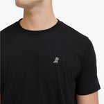 A close-up of a man wearing a black crewneck t-shirt with a small white embroidered dog logo on the left chest