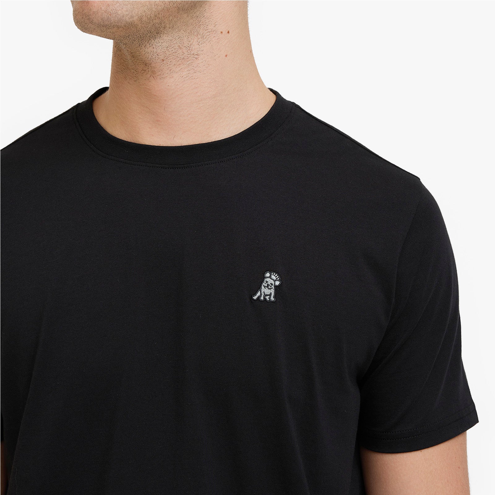 A close-up of a man wearing a black crewneck t-shirt with a small white embroidered dog logo on the left chest