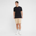 A man modeling a black T-shirt with a small white embroidered logo on the left chest, paired with beige shorts, white socks, and white sneakers