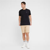 A man modeling a black T-shirt with a small white embroidered logo on the left chest, paired with beige shorts, white socks, and white sneakers