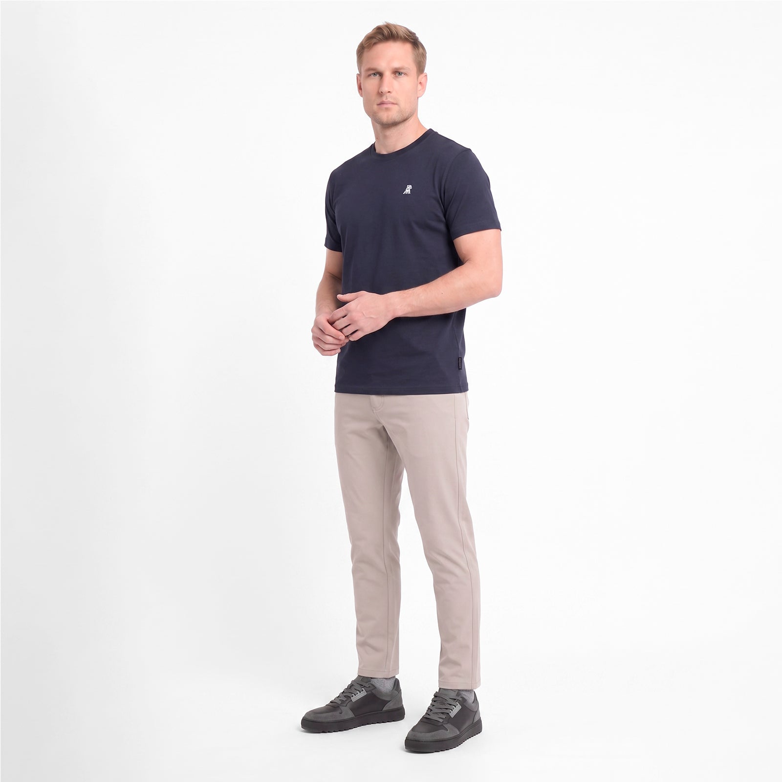A man wearing a short-sleeve crewneck t-shirt in navy blue with a small white logo on the left chest, paired with beige trousers