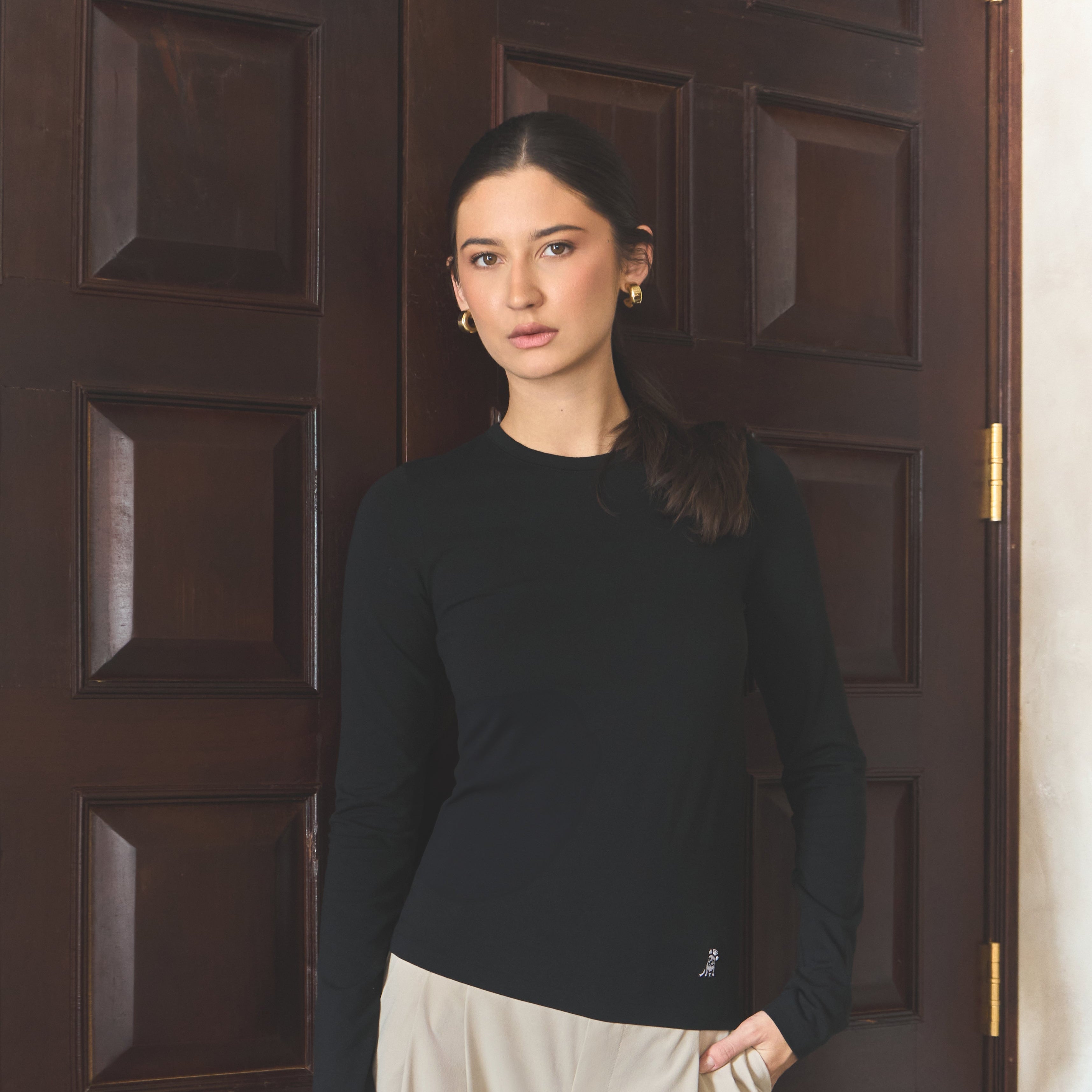 Woman wearing a black sweater and white pants standing in front of a dark wooden door.