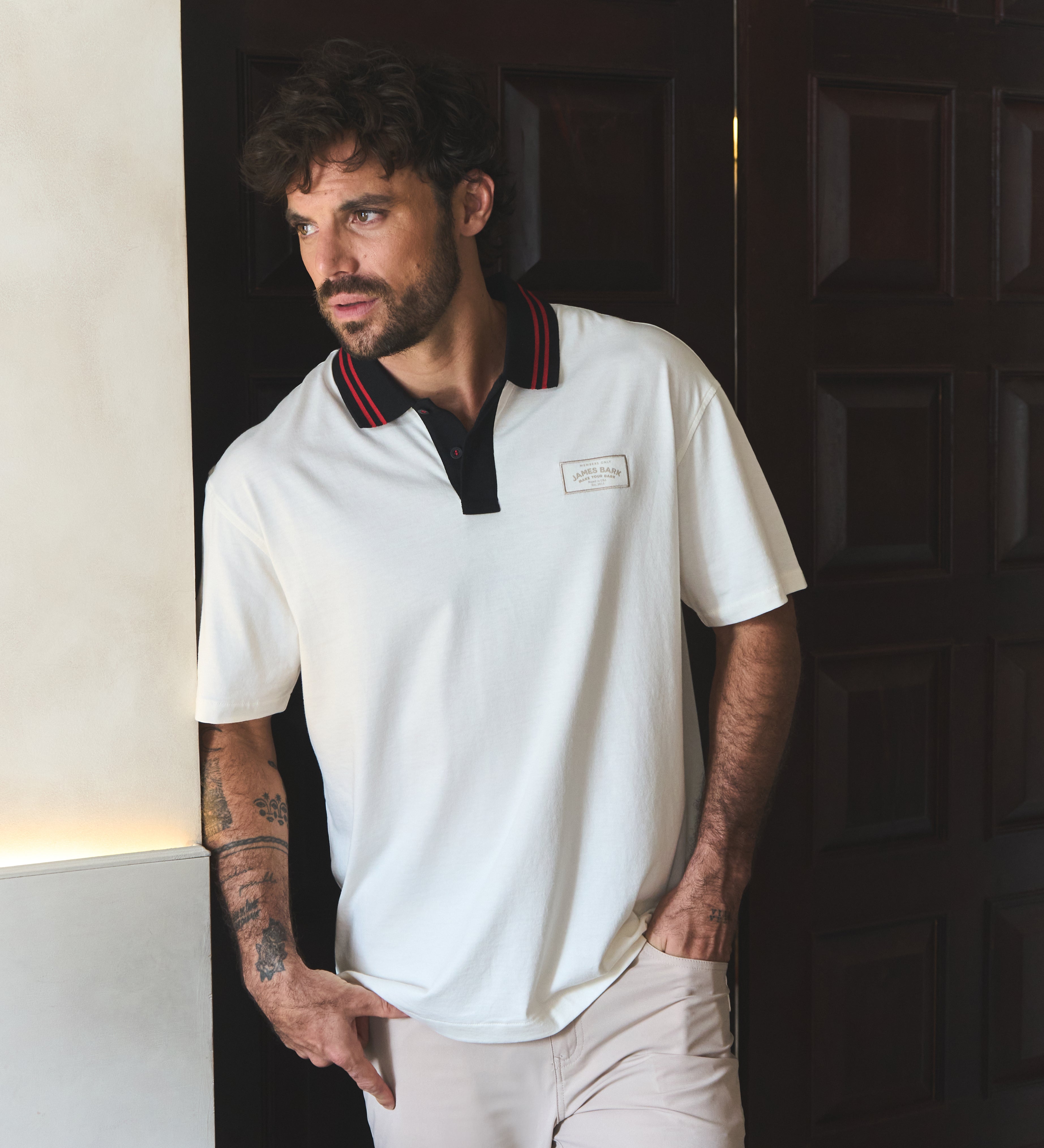 Man wearing a white polo shirt with red trim standing against a dark wooden door.