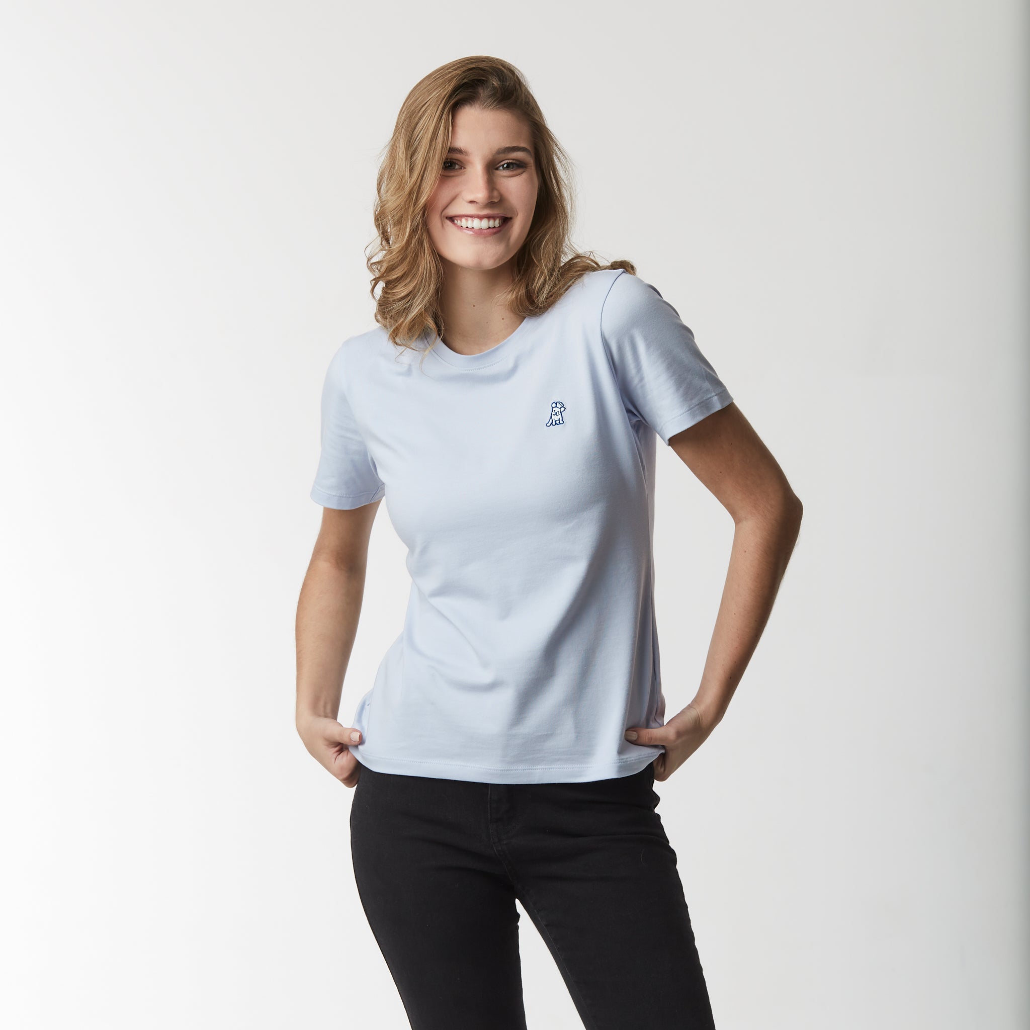 Front view of a woman wearing a light blue t-shirt paired with black pants, smiling with hands on her hips.