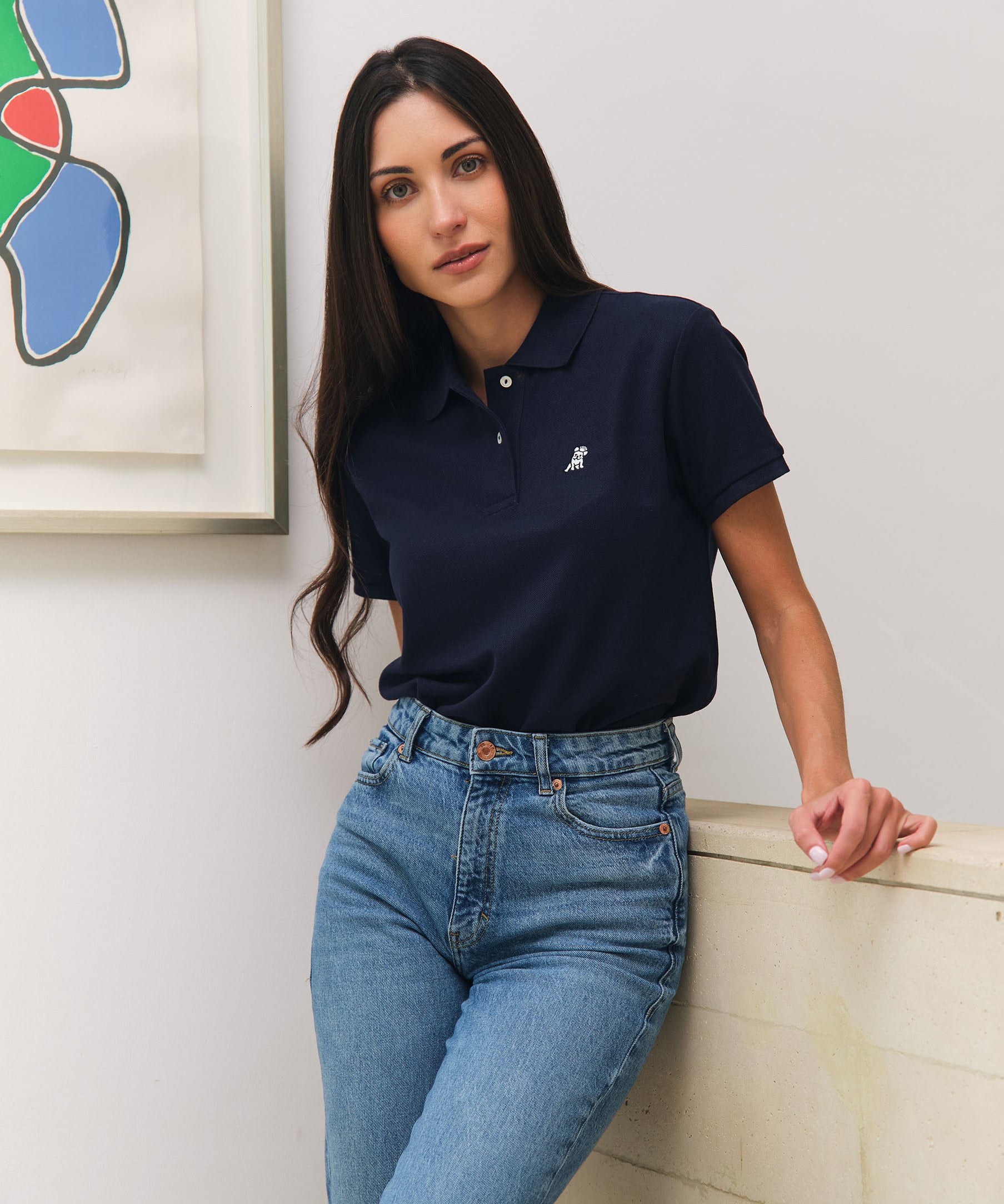 A woman wearing a navy blue polo shirt with a small white embroidered dog logo on the chest, paired with high-waisted light blue jeans. She is leaning against a light-colored wall and looking directly at the camera. There is a colorful abstract painting on the wall beside her, and the setting is clean and minimalistic.