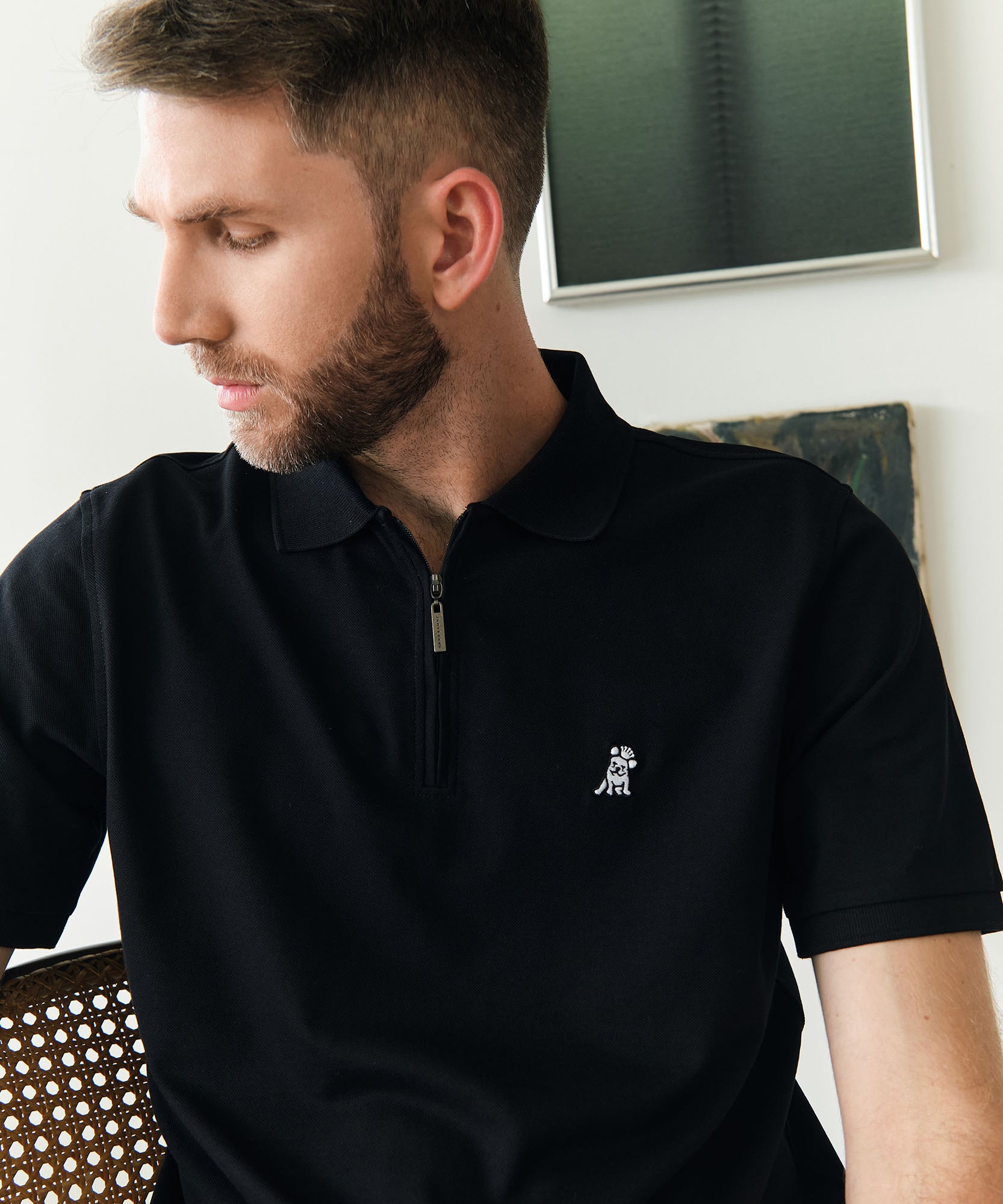 A man sitting on a chair and wearing a black polo shirt with a half-zip closure. The shirt features a small white embroidered dog logo on the chest. The man, with short brown hair and a trimmed beard, looks down to his left. In the background, there is a modern art piece and a light-colored wall.