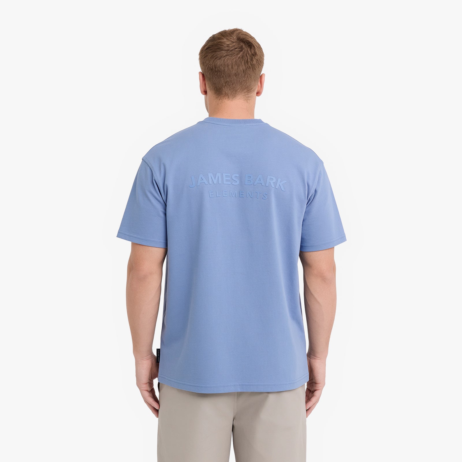 A man viewed from behind wearing a light blue t-shirt with 'JAMES BARK ELEMENTS' printed in tonal light blue on the back, paired with beige trousers