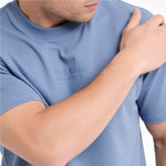 A close-up of a man wearing a light blue t-shirt with tonal embossed text on the chest reading 'JAMES BARK ELEMENTS'.