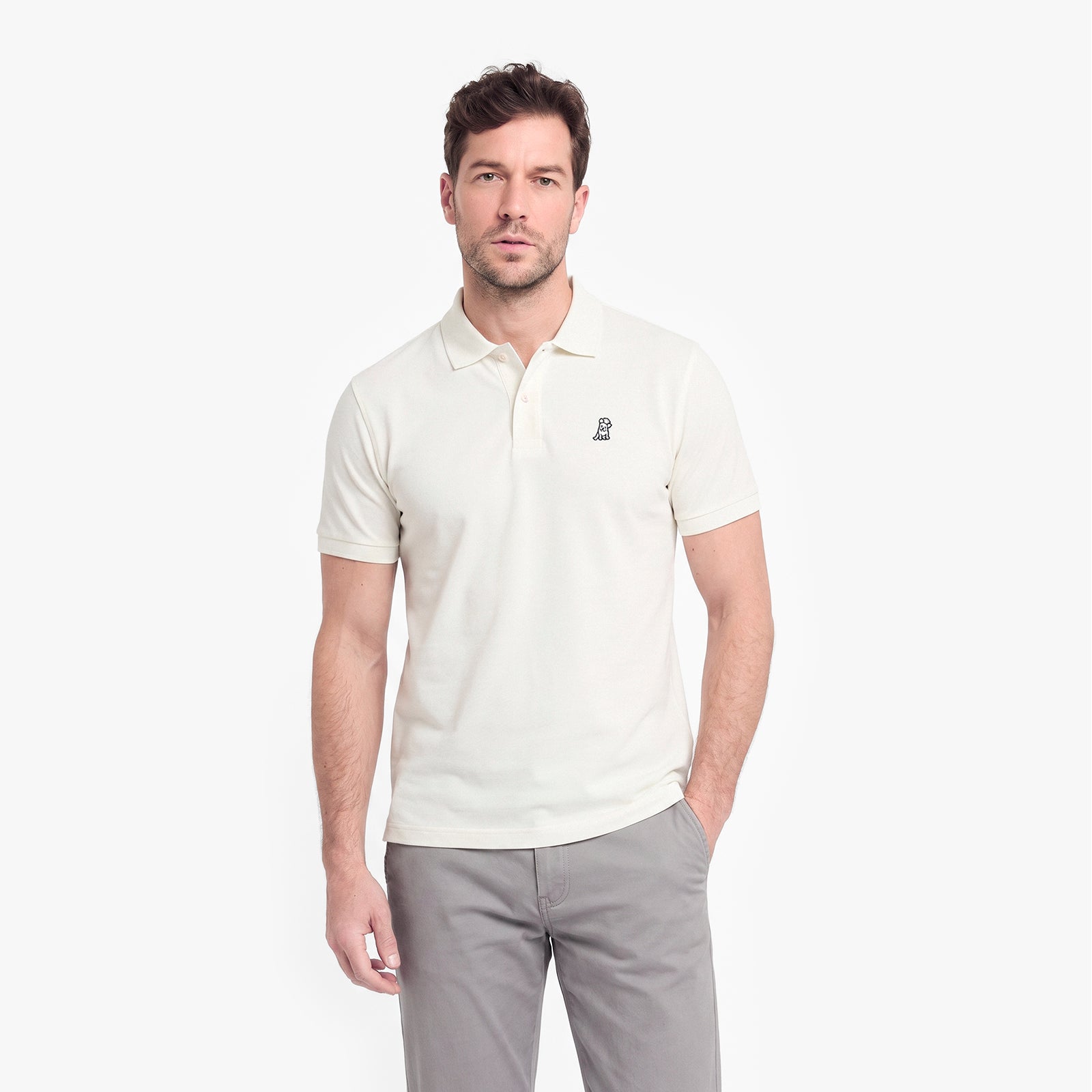 A man wearing a short-sleeve polo shirt in off-white with a small dark embroidered logo on the left chest, paired with gray trousers