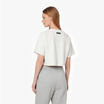 A woman shown from the back wearing a cropped white T-shirt with a small black rectangular patch on the upper back, paired with light gray sweatpants