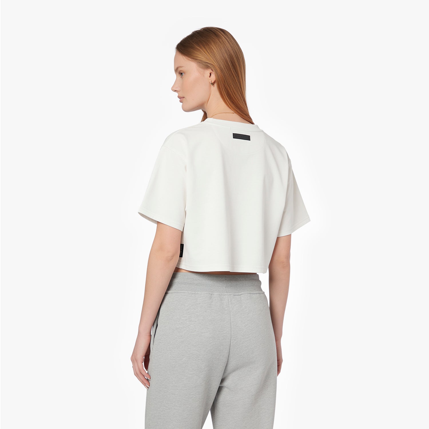 A woman shown from the back wearing a cropped white T-shirt with a small black rectangular patch on the upper back, paired with light gray sweatpants
