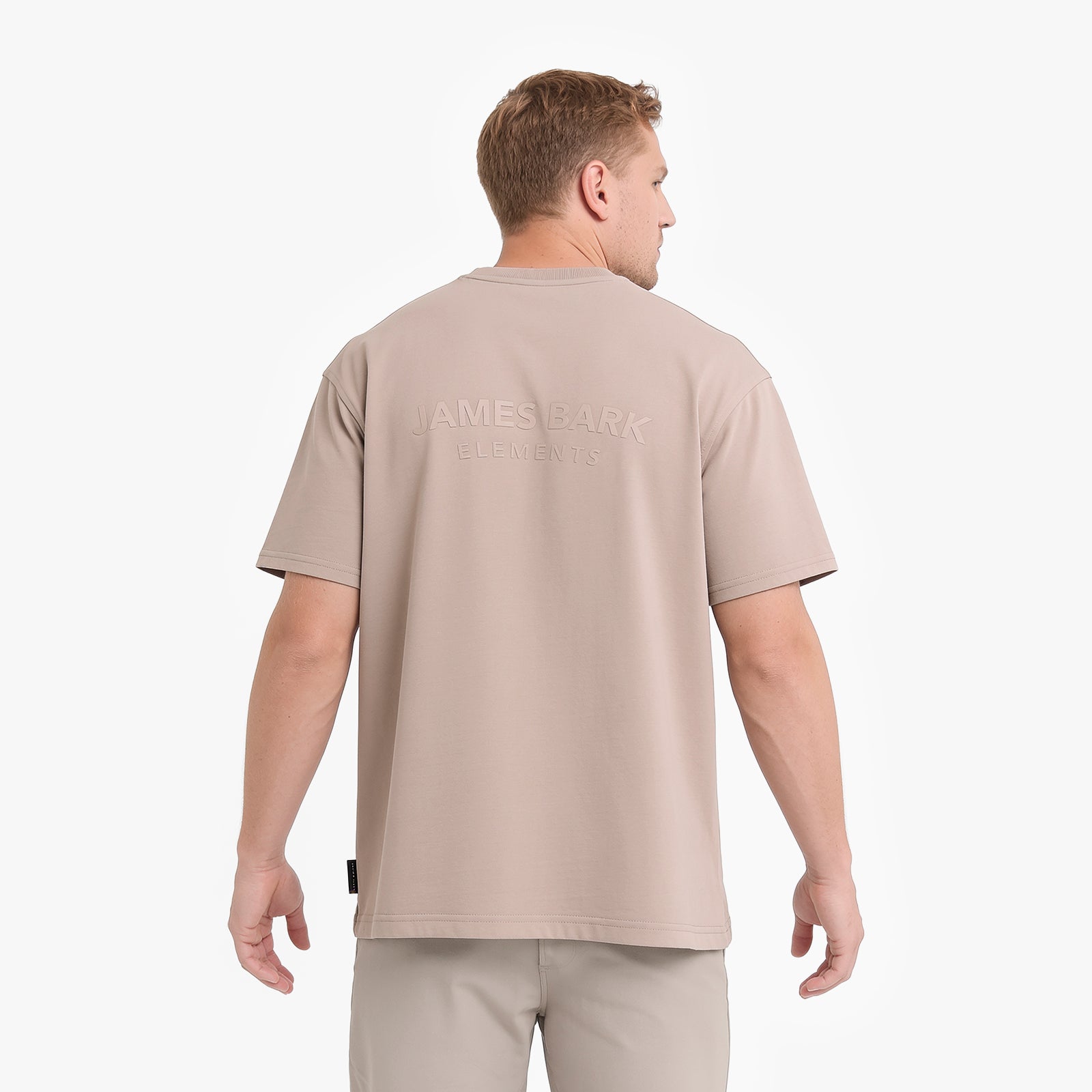 A man viewed from the back wearing a light beige short-sleeve t-shirt with 'JAMES BARK ELEMENTS' printed on the upper back, paired with matching beige trousers