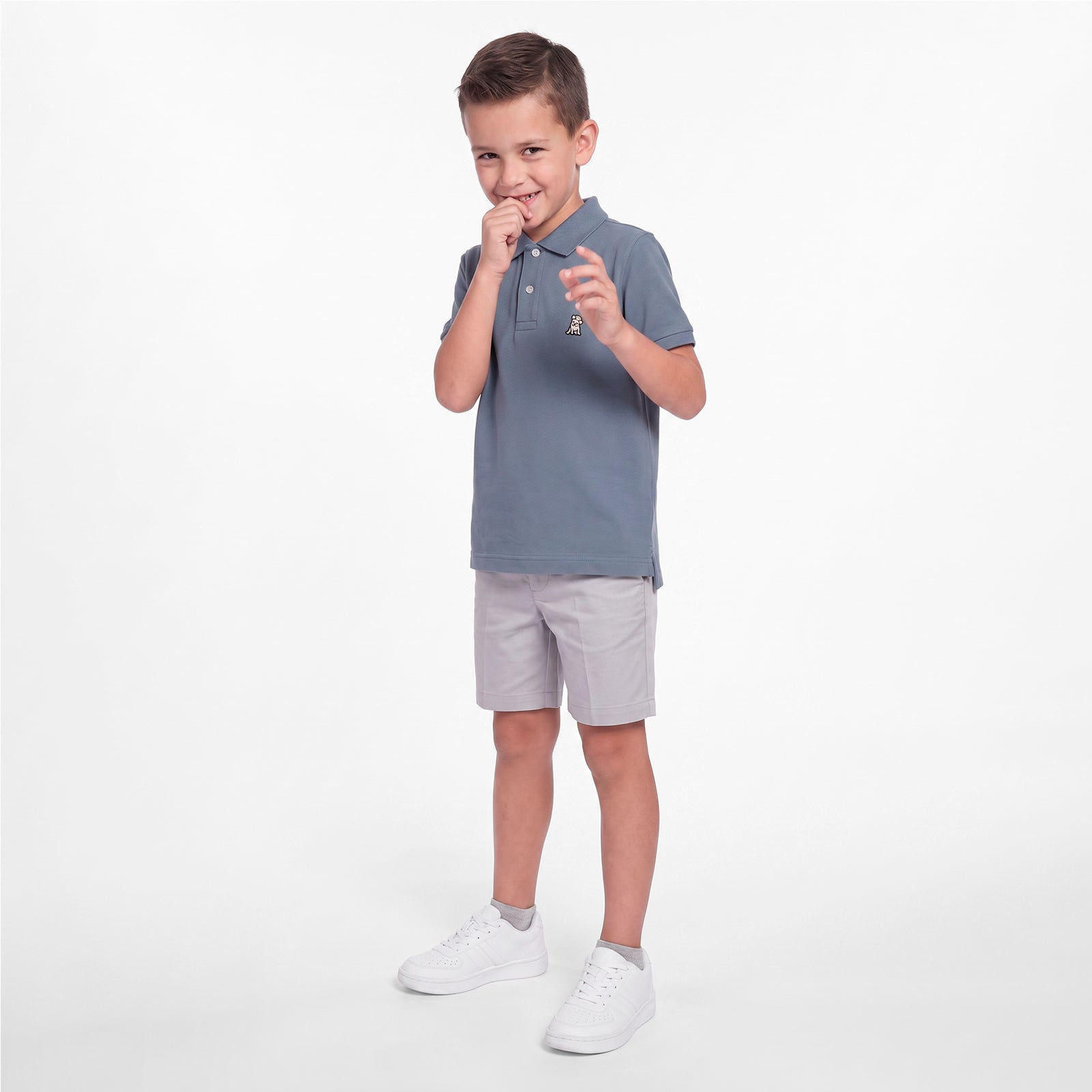 A young boy modeling a slate blue polo shirt, light gray shorts, and white sneakers