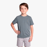 A young boy wearing a slate gray T-shirt with a small embroidered logo on the left chest, paired with light gray shorts