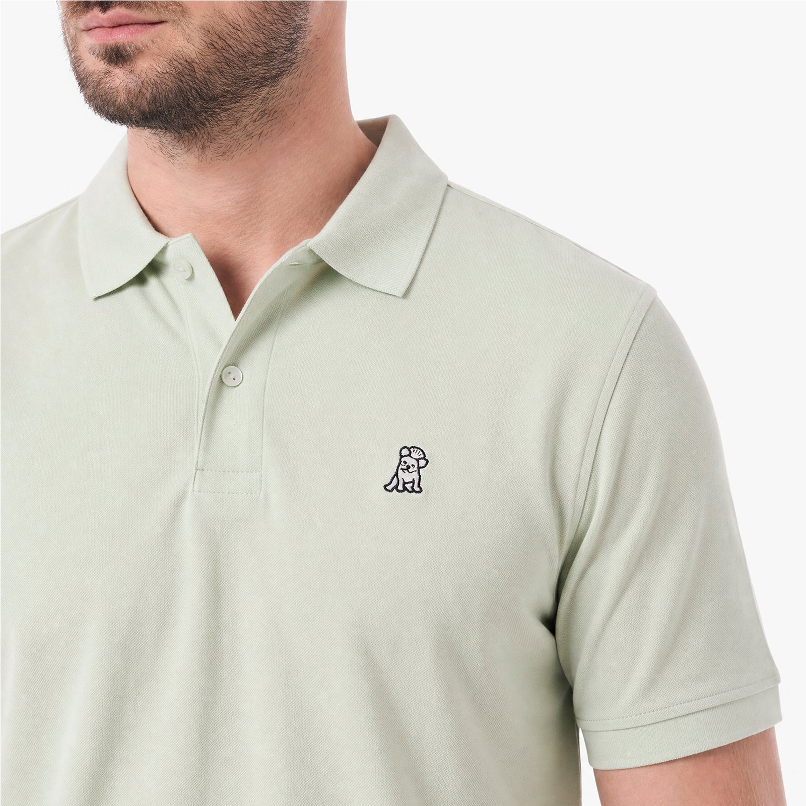A close-up of a man wearing a light sage green polo shirt with a small embroidered dog logo on the left chest