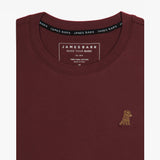 A close-up of the neckline and collar tag of the maroon t-shirt, showing part of the chest area with a small embroidered graphic of a dog.