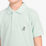 A close-up of a child wearing a pale mint green polo shirt with a small embroidered dog logo on the left chest
