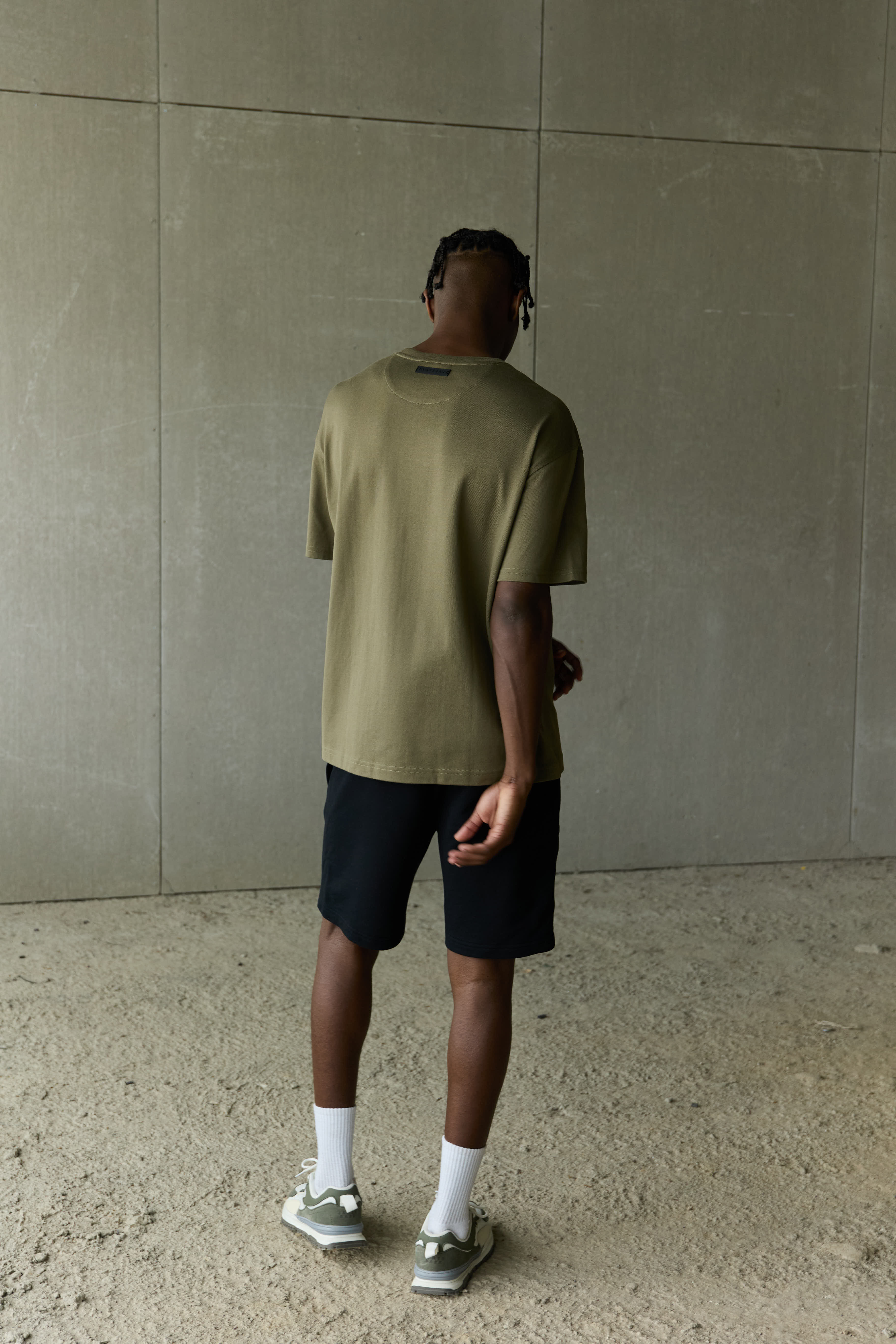 Man standing with his back to the camera, wearing an olive green T-shirt, black shorts, white socks, and green sneakers, against a textured concrete wall background.