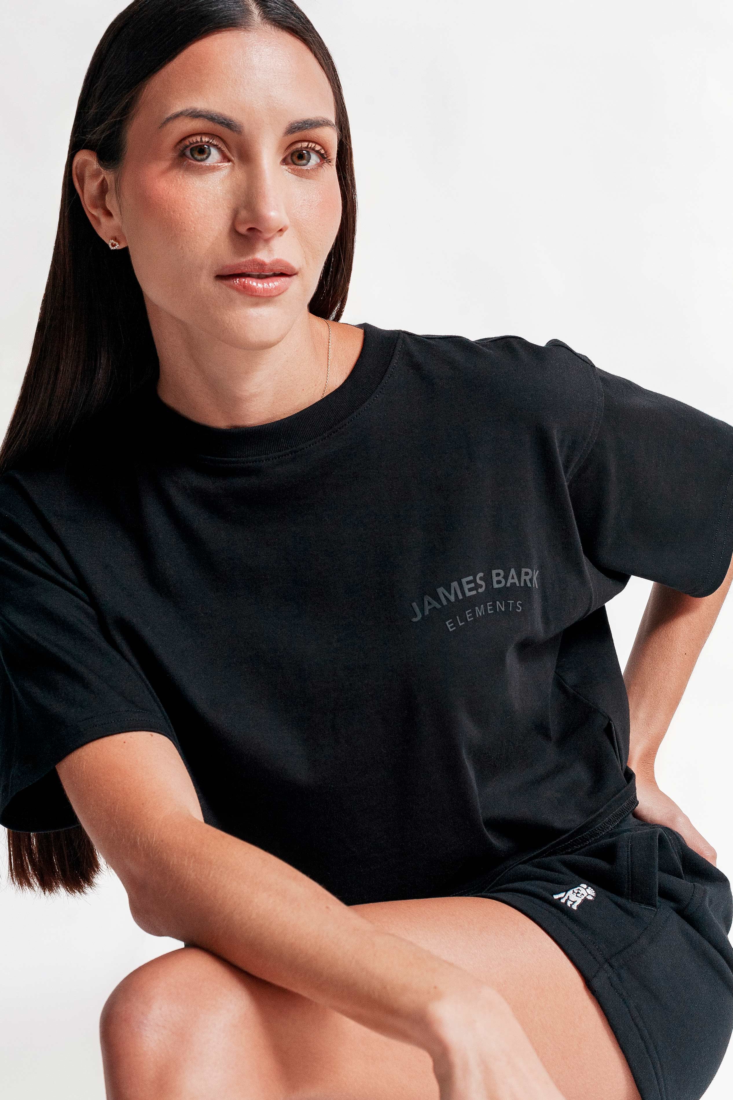 A woman with sleek dark hair and a confident expression wears a black oversized T-shirt with 'JAMES BARK ELEMENTS' printed on the upper left chest and black shorts featuring an embroidered dog logo. She sits casually against a plain white background.