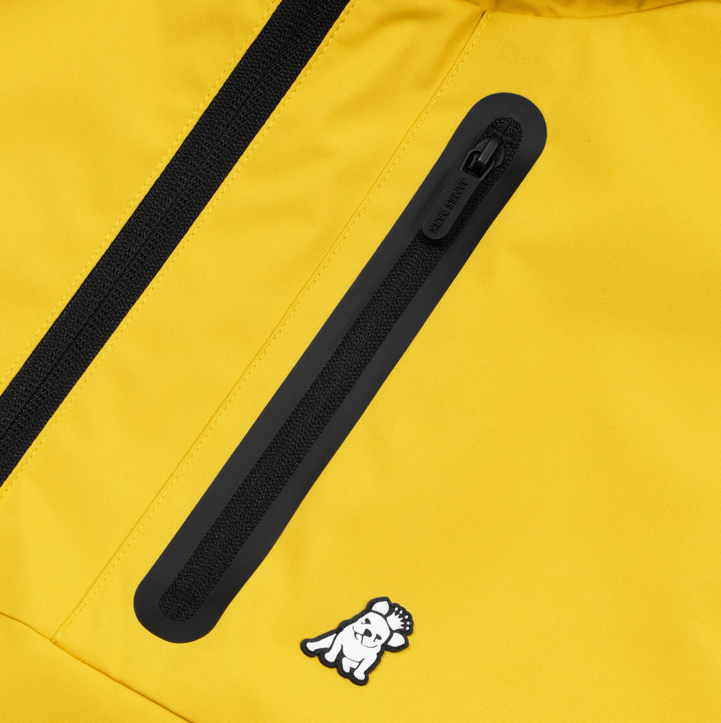 A close-up of the bright yellow jacket with a black waterproof zipper detail along the chest. The jacket features a small, embroidered white dog mascot wearing a crown, adding a playful element to the functional, sporty design. The black zipper is sleek and contrasts against the vibrant yellow fabric, which looks smooth and durable.