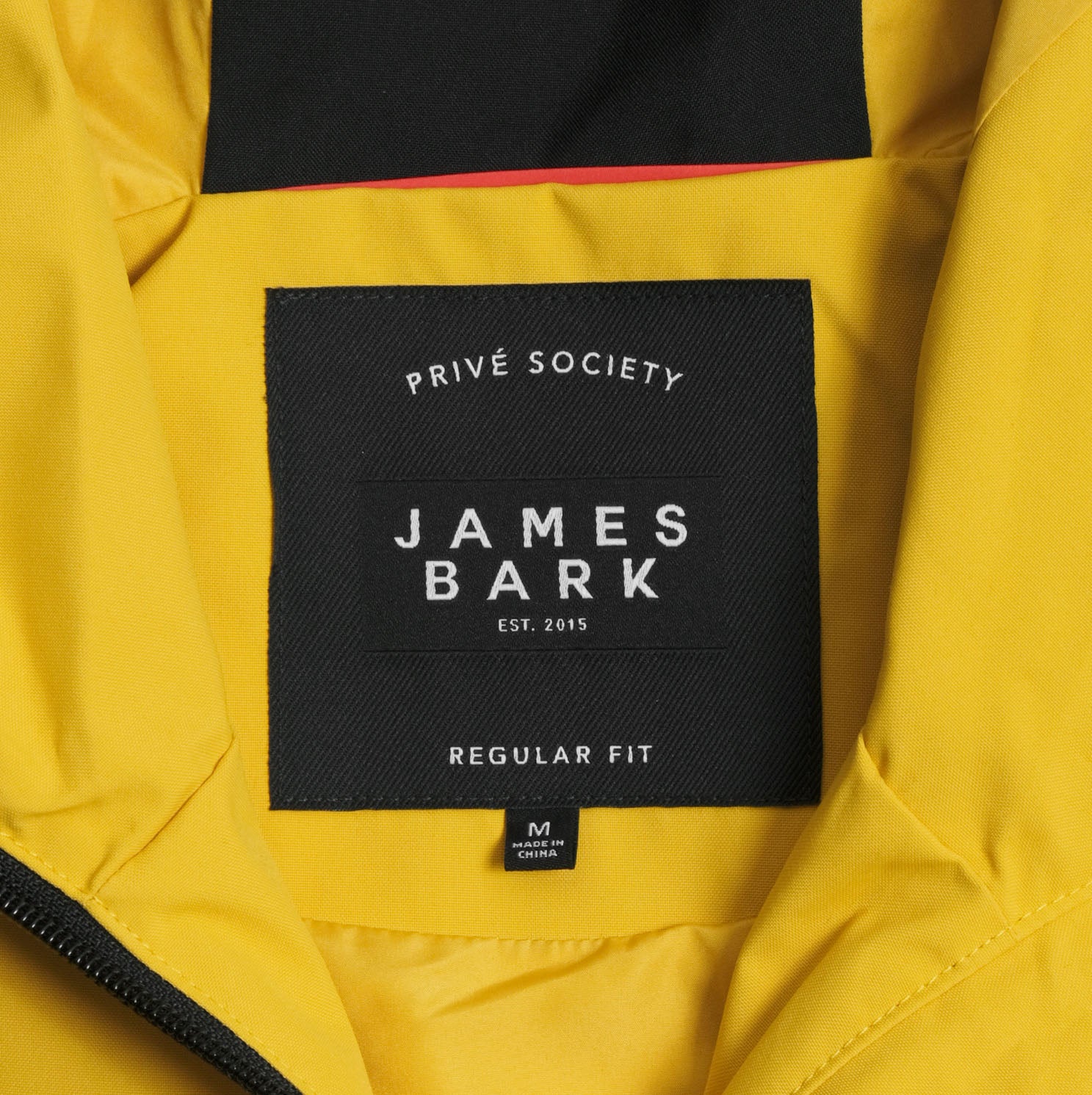 Close-up image of the inner label of a yellow James Bark jacket. The label reads 'PRIVÉ SOCIETY' at the top, 'JAMES BARK EST. 2015' in the center, and 'REGULAR FIT' at the bottom. Below is a small tag indicating the jacket is 'Made in China.' The label is black with white text, and the yellow fabric of the jacket surrounds the label.