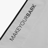 Close-up of a light gray fabric featuring the words 'MAKEYOURBARK' printed in bold, black lettering along the side of a black zipper. The text stands out clearly against the light background, maintaining a sleek and minimalist design.