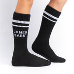 Black mid-calf socks with two white stripes at the top and the text 'JAMES BARK' in white on the ankle, shown worn on a person’s feet.




