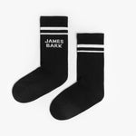 A pair of black mid-calf socks with two white stripes at the top and the text 'JAMES BARK' printed in white near the ankle. The socks are laid flat on a white surface.