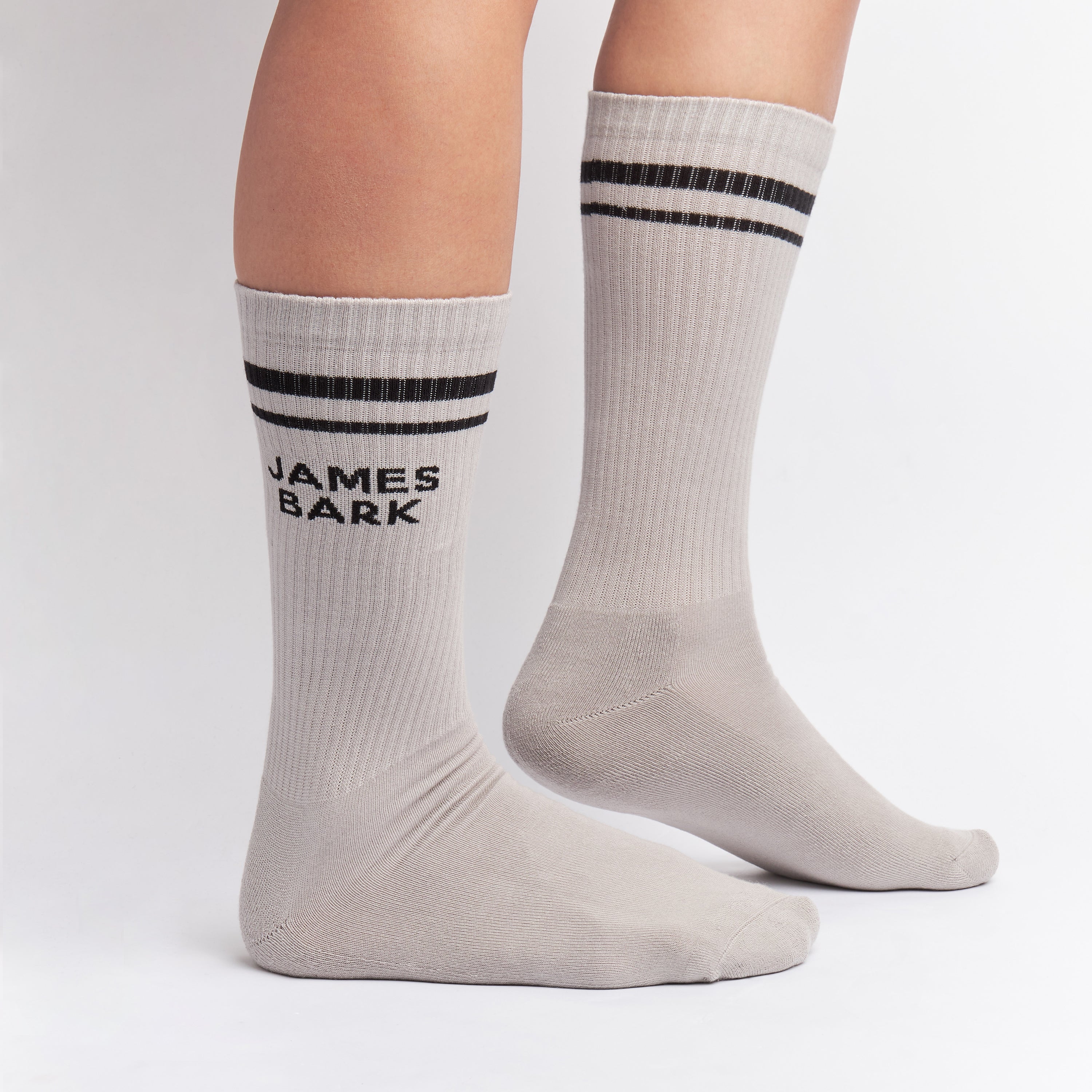 Light gray mid-calf socks with two black stripes at the top and the text 'JAMES BARK' in black on the ankle, shown worn on a person’s feet.