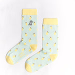 A pair of light blue mid-calf socks with a yellow cuff, heel, and toe, featuring a pattern of lemons and a small dog graphic near the ankle. The text 'JAMES BARK' is printed in yellow near the toe. The socks are laid flat on a white surface.