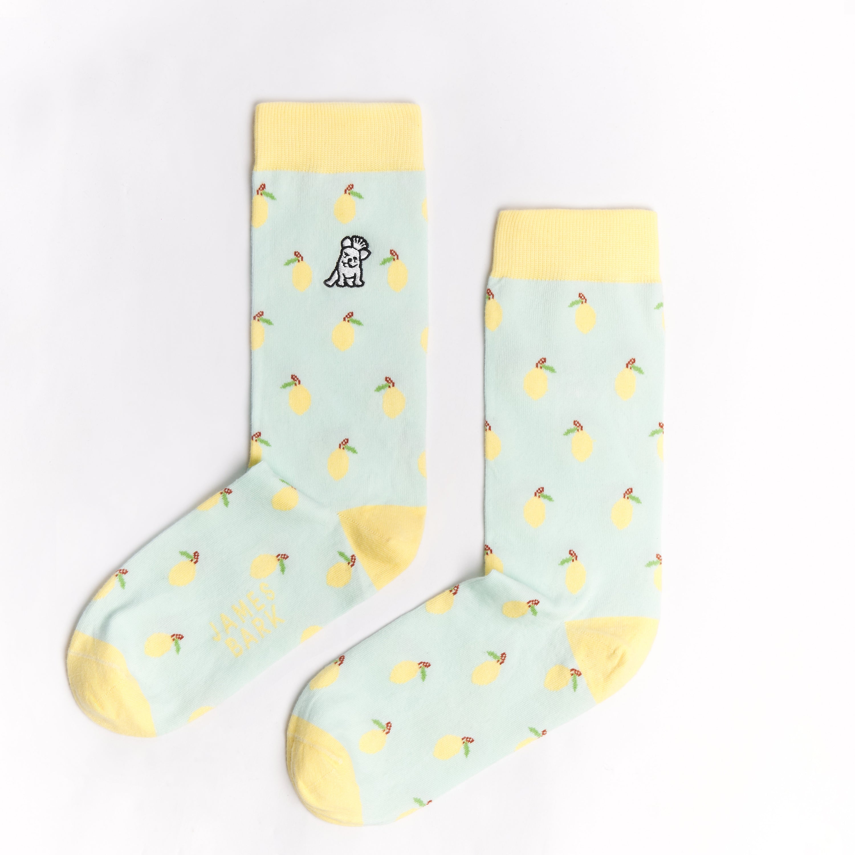 A pair of light blue mid-calf socks with a yellow cuff, heel, and toe, featuring a pattern of lemons and a small dog graphic near the ankle. The text 'JAMES BARK' is printed in yellow near the toe. The socks are laid flat on a white surface.