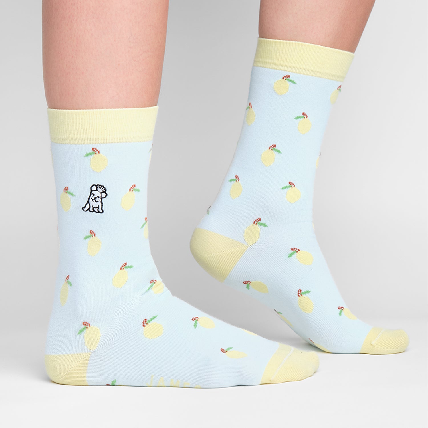 A close-up of a person's legs wearing light blue socks with a lemon pattern, featuring small yellow lemons with green leaves. The socks have a light yellow cuff, heel, and toe, and a small embroidered logo near the top of the socks.