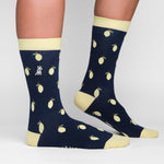 A close-up of a person's legs wearing navy blue socks with a lemon pattern, featuring small yellow lemons with green leaves. The socks have a light yellow cuff, heel, and toe, and a small embroidered logo near the top of the socks.
