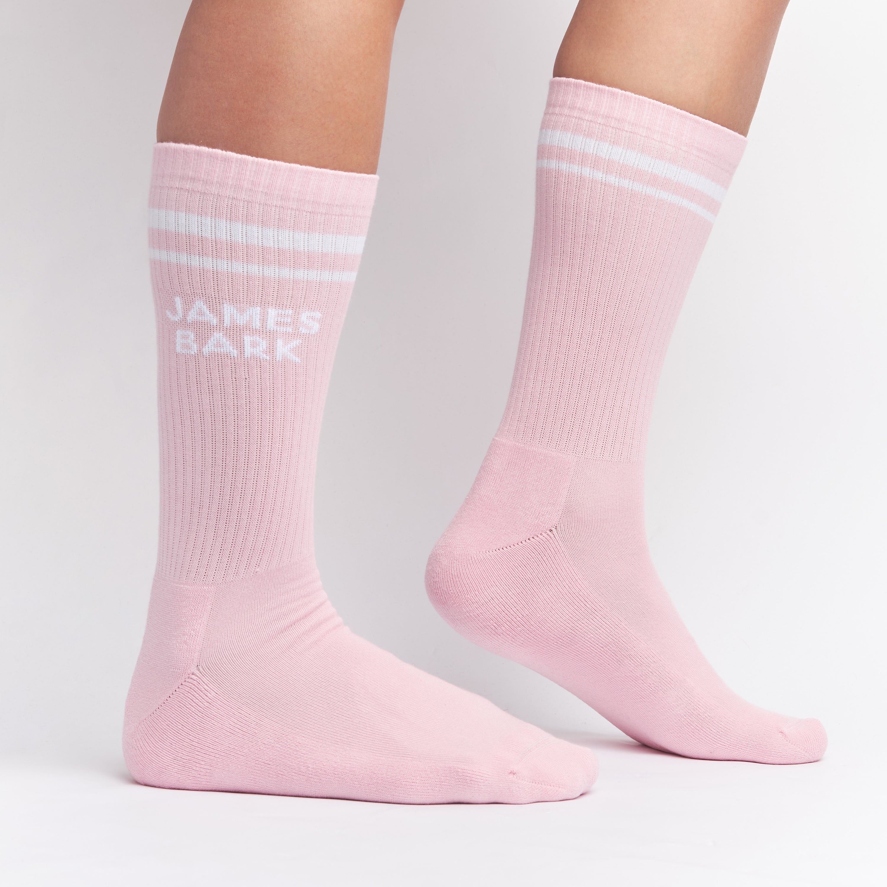 Pink mid-calf socks with two white stripes at the top and the text 'JAMES BARK' in white on the ankle, shown worn on a person’s feet.