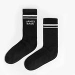 A pair of black mid-calf socks with two white stripes at the top and the text 'JAMES BARK' printed in white near the ankle. The socks are laid flat on a white surface.