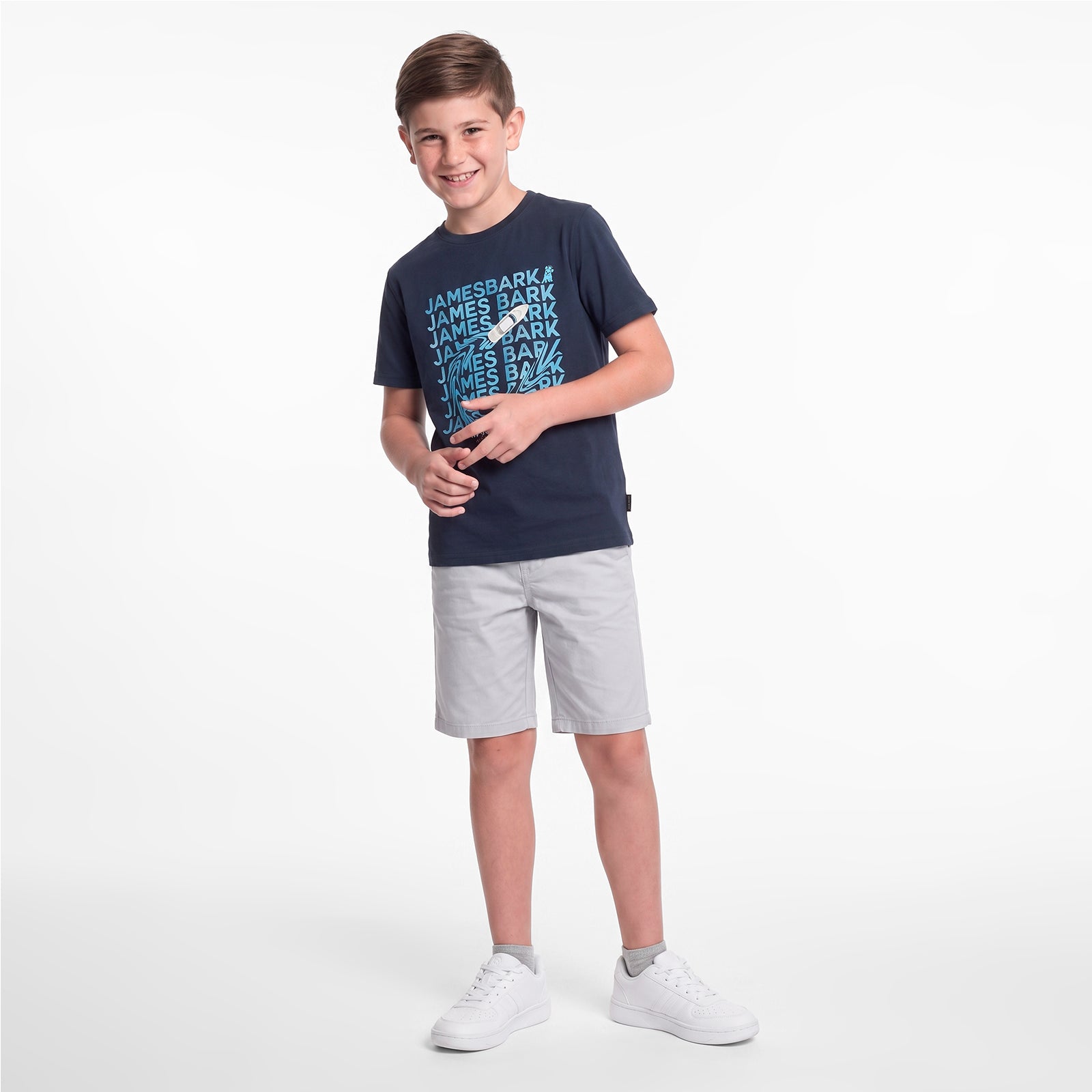 A young boy wearing a navy blue T-shirt with a light blue and white 'JAMES BARK' graphic, paired with light gray shorts and white sneakers