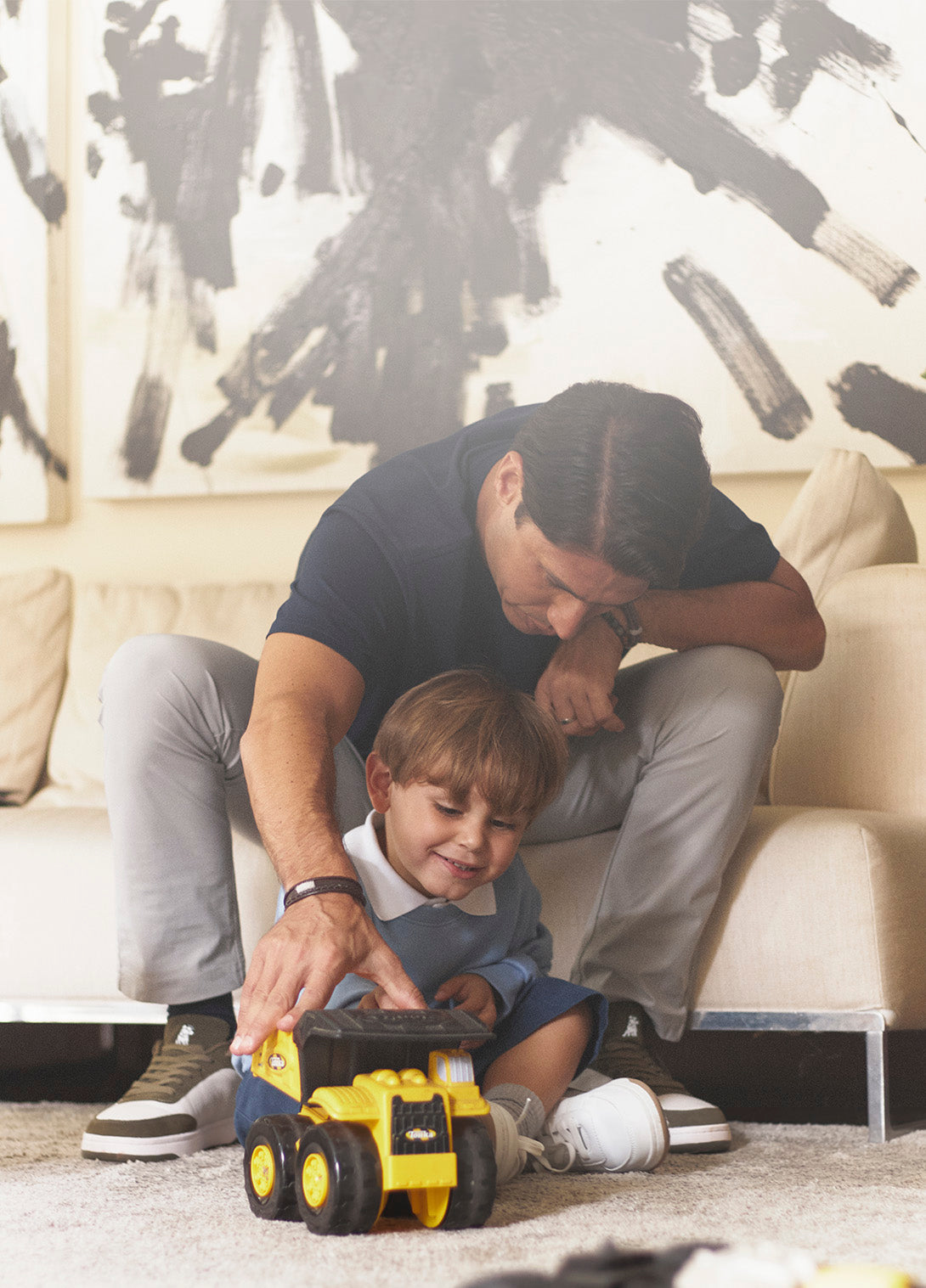 A dad kneels next to his smiling son, who wears a pastel blue long-sleeve polo with a white collar. The pair play with a yellow toy truck on a cozy beige carpet in a modern, art-filled living room