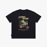A black T-shirt shown flat, featuring a large graphic print on the back with the text 'JAMES BARK' and 'Luxury by the Coast'. The graphic depicts a sunset beach scene with a pool and palm trees.