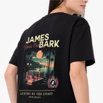 A woman wearing a black oversized T-shirt. The shirt features a large graphic print with the text 'JAMES BARK' and 'Luxury by the Coast' in cream and pink, along with a tropical beach scene illustration.