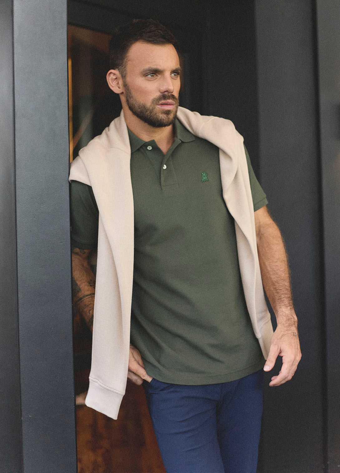 Stylish man in an olive green polo shirt and beige sweatshirt draped over his shoulders, posing confidently outside a contemporary glass-paneled building with indoor lighting and plants visible in the background