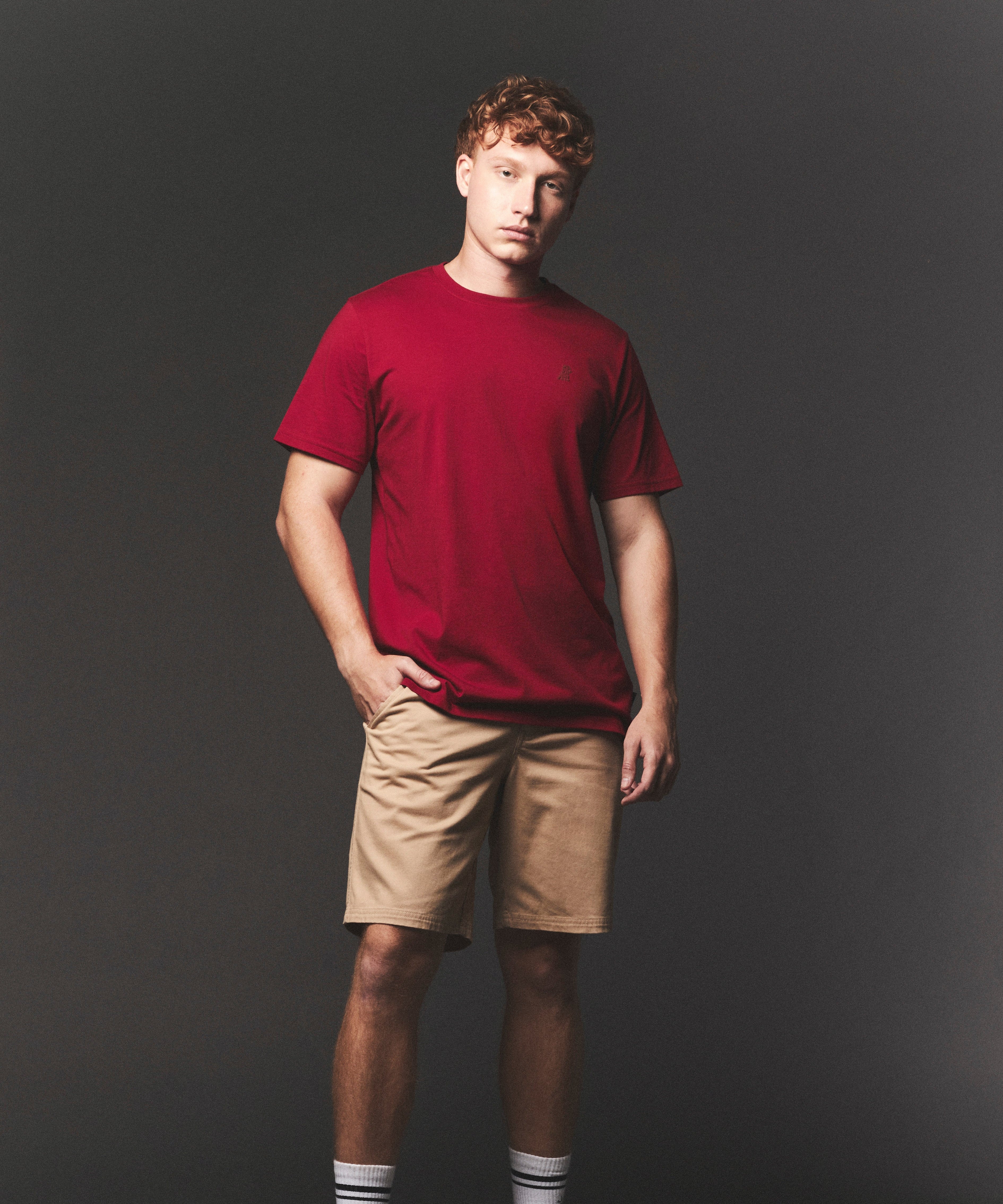 Man wearing a red t-shirt and beige shorts against a dark background