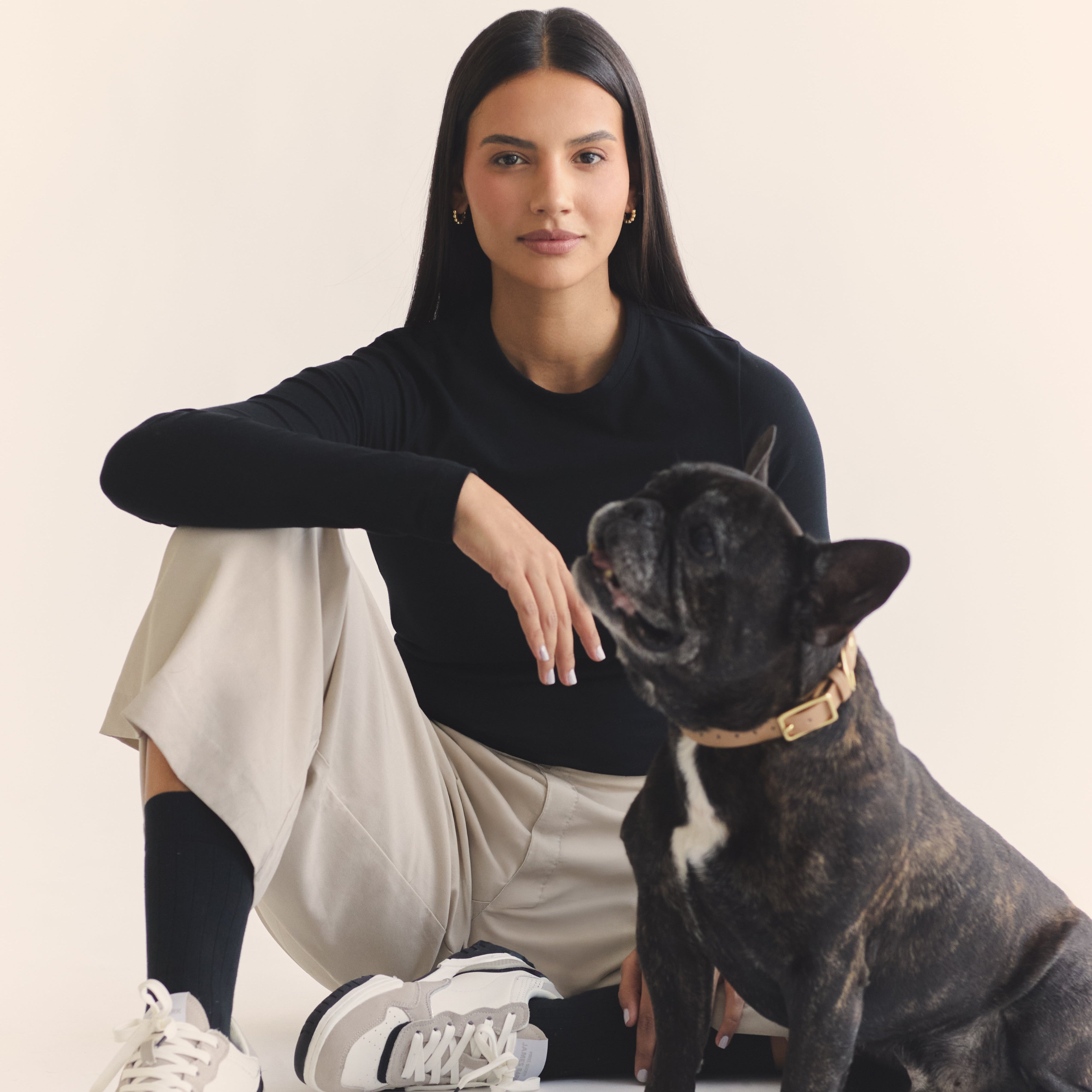 Woman wearing a black long sleeve t-shirt and beige pants, next to a french bulldog on a gray background