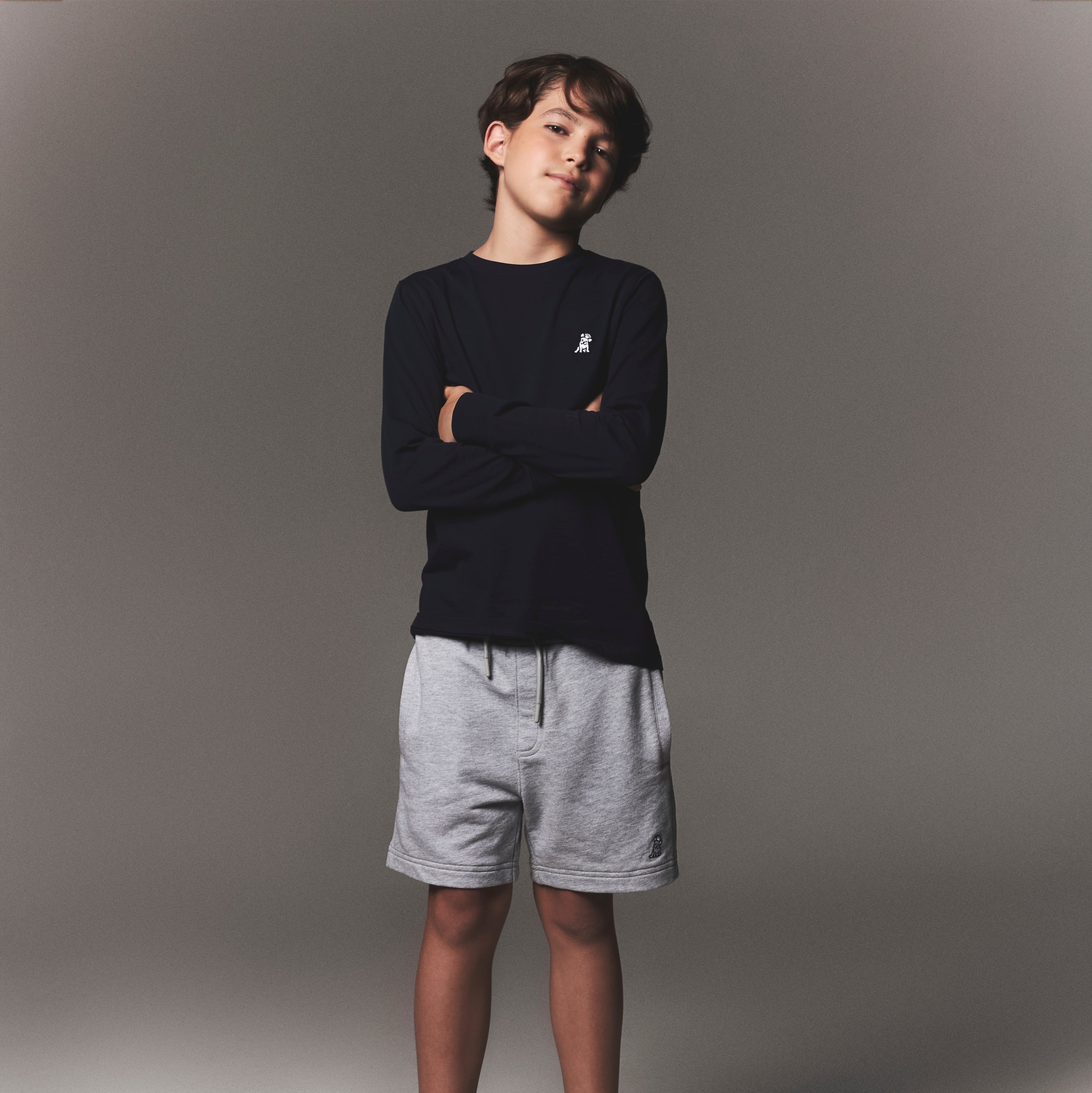 Child wearing a black long-sleeve shirt, gray shorts, and white sneakers on a gray background